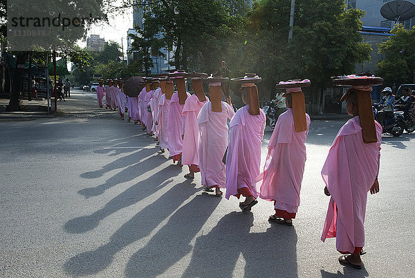 Lange Reihe buddhistischer Nonnen in rosafarbenen Gewändern  Köpfe bedeckt  Almosenschalen auf dem Kopf  beim Überqueren der Hauptstraße in Mandalay  Myanmar (Burma)  Asien