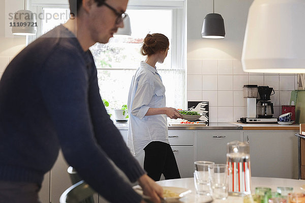 Mann arrangiert den Esstisch  während die Frau das Essen in der Küche zubereitet.