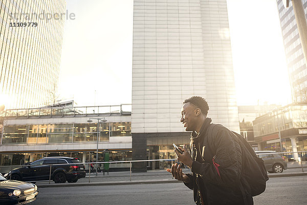 Lächelnder Teenager gestikuliert  während er Telefon gegen Gebäude in der Stadt hält.