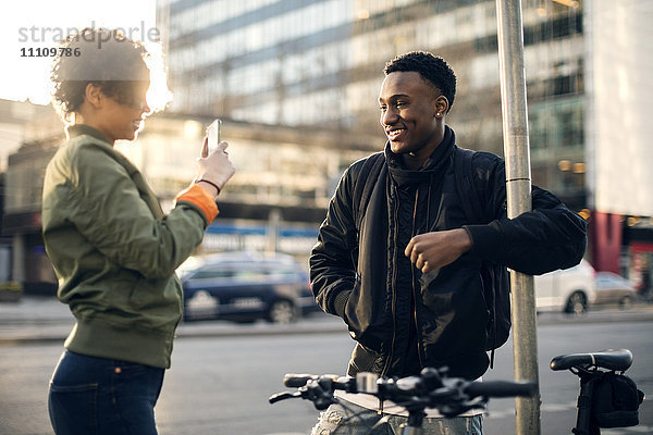 Seitenansicht eines Teenagers  der einen Freund über ein Smartphone fotografiert  während er an der Stadtstraße steht.