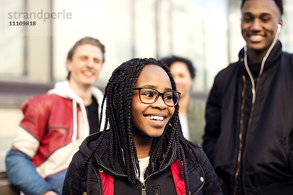 Lächelnder Teenager mit geflochtenem Haar  der eine Brille trägt und gegen Freunde in der Stadt steht.