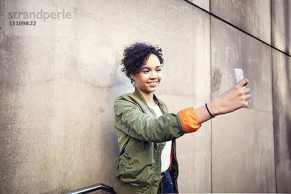 Lächelndes Teenagermädchen nimmt Selfie durchs Handy  während es an der verwitterten Wand steht.