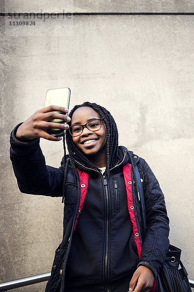 Niedriger Blickwinkel eines lächelnden Teenagers  der Selfie durch ein Smartphone gegen die Wand nimmt.