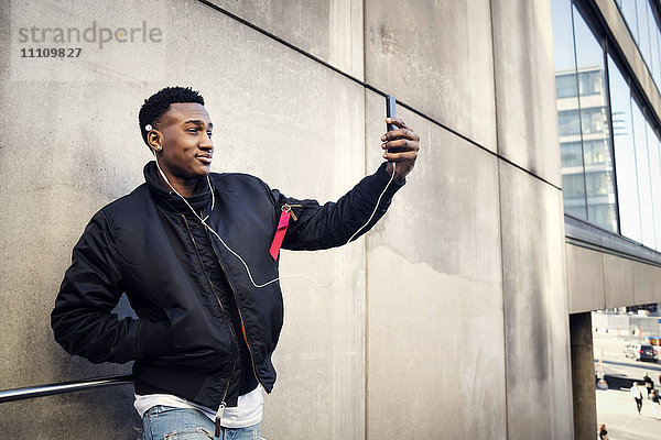 Lächelnder Teenager nimmt Selfie durch das Smartphone  während er Musik gegen die Wand hört.