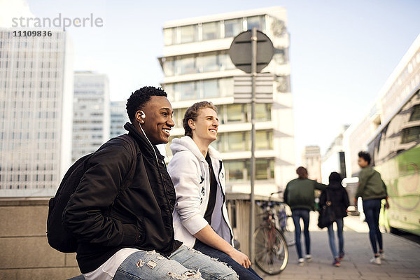 Fröhliche Teenager  die sich auf dem Geländer ausruhen  während Freunde in der Stadt auf dem Fußweg spazieren gehen