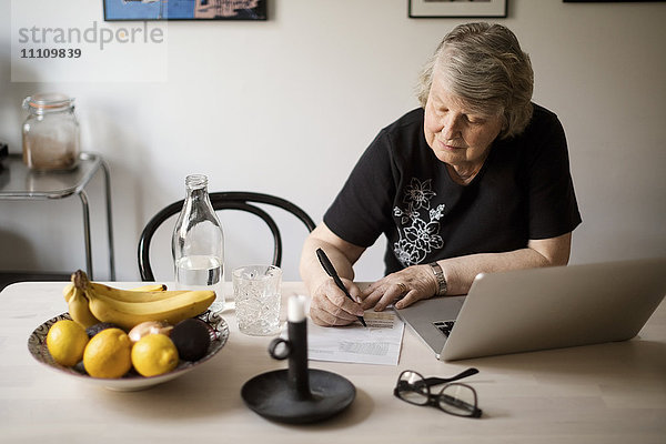 Seniorenfrau  die nach Rechnungen sucht  während sie den Laptop am Tisch benutzt.