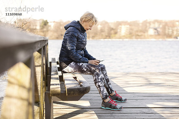 Seniorenfrau in Sportbekleidung mit Telefon auf der Bank am See