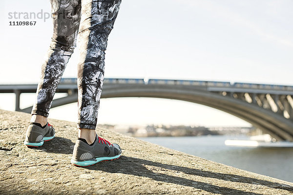 Niedriger Abschnitt der Frau mit Sportschuhen  die auf einem Felsen an einer Brücke steht.