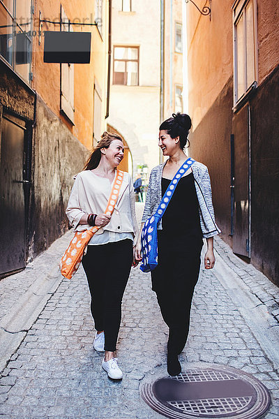 Volle Länge von Freunden  die sich unterhalten  während sie durch die Gasse inmitten von Gebäuden in der Stadt spazieren gehen.