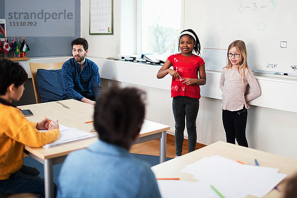 Schülerinnen stehen gegen Whiteboard und Lehrerin im Klassenzimmer