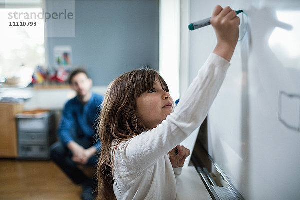 Selbstbewusstes Mädchen malt an der Tafel  während die Lehrerin im Hintergrund sitzt.