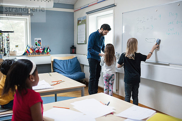 Mädchen im Gespräch mit dem Lehrer  während der Schüler auf einer Tafel im Klassenzimmer malt.