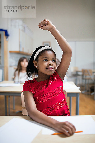 Konzentriertes Mädchen mit erhobener Hand am Schreibtisch im Klassenzimmer sitzend