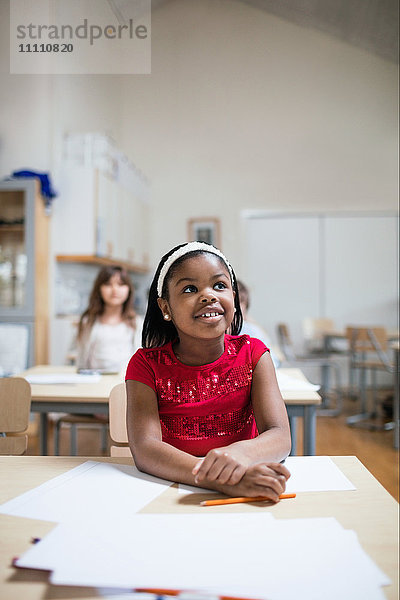 Lächelndes Mädchen  das am Schreibtisch im Klassenzimmer sitzt und zuhört.