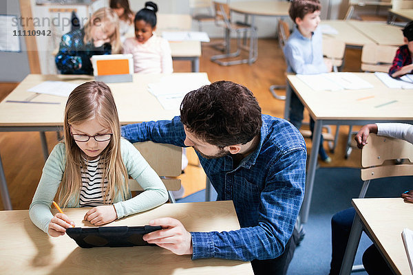 Hochwinkelansicht des Lehrers  der die Schülerin bei der Verwendung des digitalen Tabletts im Klassenzimmer unterstützt.