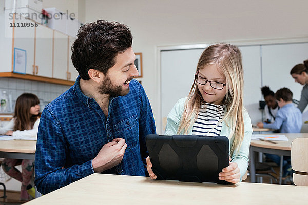 Lächelnde Lehrerin schaut Mädchen mit digitalem Tablett am Schreibtisch an