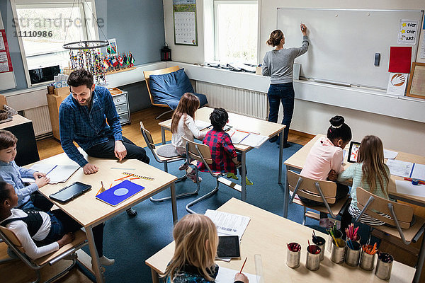 Hochwinkelansicht der Lehrerin beim Schreiben an Bord  während der Lehrer mit den Schülern am Schreibtisch im Klassenzimmer sitzt.