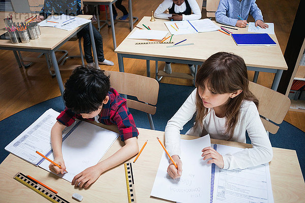 Hochwinkelansicht von Schülern  die auf Papier schreiben  während sie im Klassenzimmer am Schreibtisch sitzen.