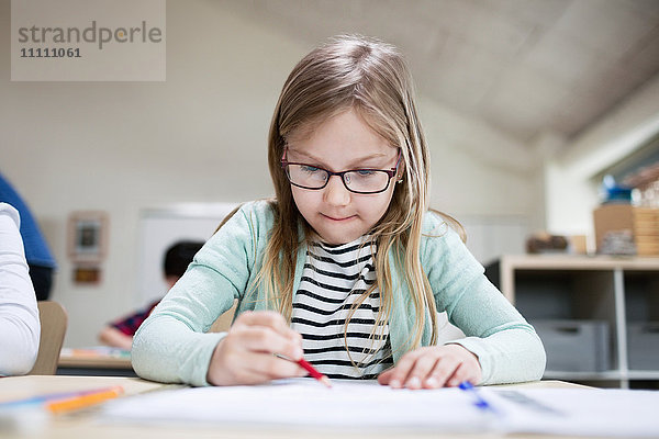 Konzentriertes Mädchen mit Brille beim Schreiben am Klassenzimmer-Schreibtisch in der Schule