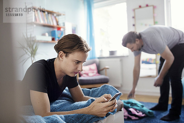 Mann  der ein Smartphone benutzt  während Vater zu Hause Kleider auswählt  die auf den Teppich gefallen sind.