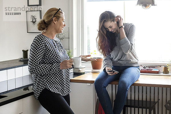 Mutter im Gespräch mit glücklicher Tochter über Smartphone in der Küche