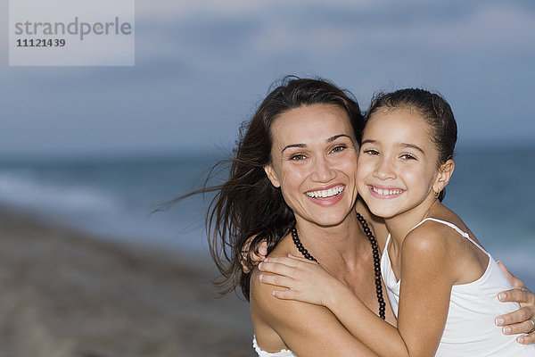 Hispanische Mutter und Tochter umarmen sich am Strand