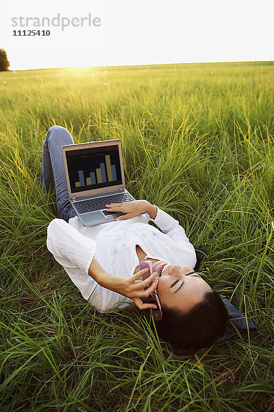 Asiatische Frau liegt auf einer Wiese und benutzt Laptop und Handy