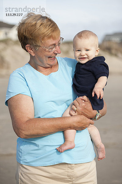 Lächelnde kaukasische Großmutter hält ihren Enkel am Strand