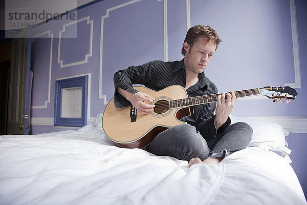 Kaukasischer Mann sitzt auf dem Bett und spielt Gitarre