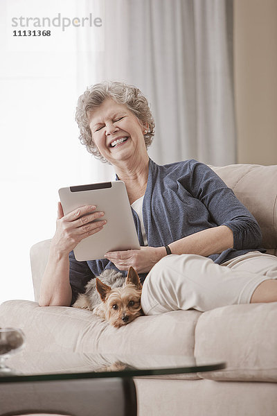 Kaukasische Frau sitzt auf dem Sofa und benutzt ein digitales Tablet mit einem Hund