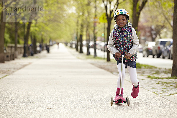 Gemischtrassiges Mädchen fährt auf einem Roller auf dem Bürgersteig