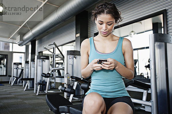 Gemischtrassige Frau  die im Fitnessstudio eine SMS auf ihrem Handy schreibt