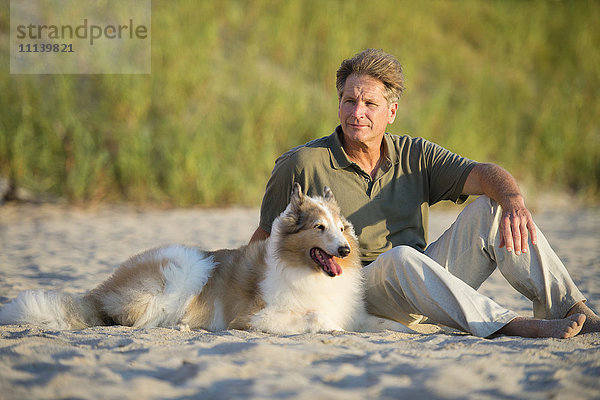 Kaukasischer Mann sitzt mit Hund am Strand