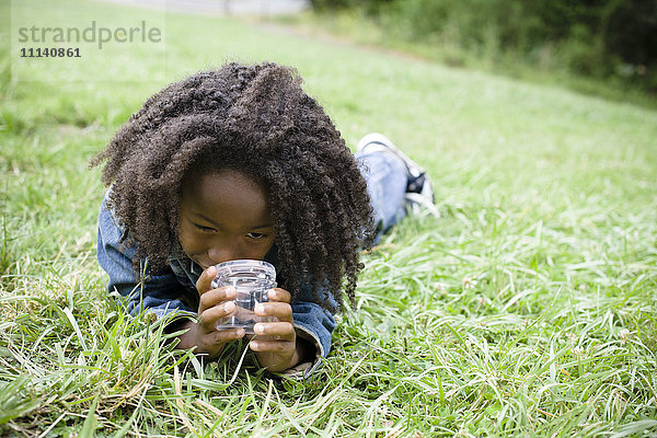 Afroamerikanischer Junge liegt im Gras und schaut in ein Glas