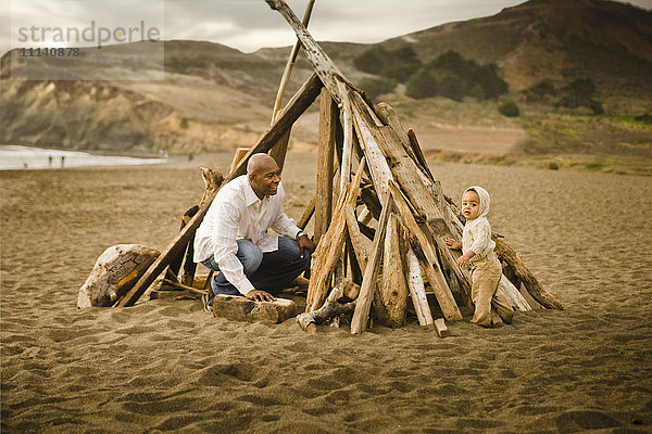 Vater und kleiner Sohn sitzen unter einem Tipi aus Treibholz am Strand