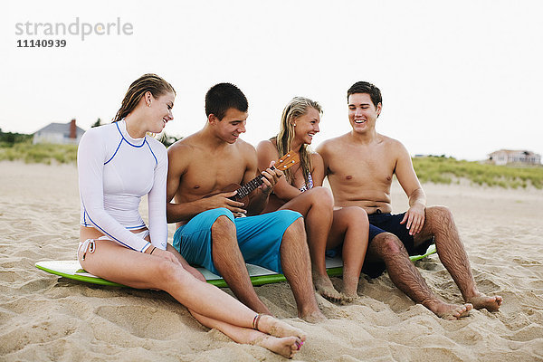 Teenager sitzen auf einem Surfbrett und spielen Ukulele am Strand