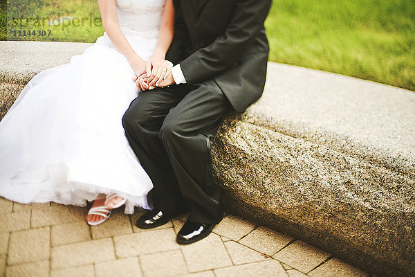 Braut und Bräutigam halten sich auf einer Steinbank an den Händen