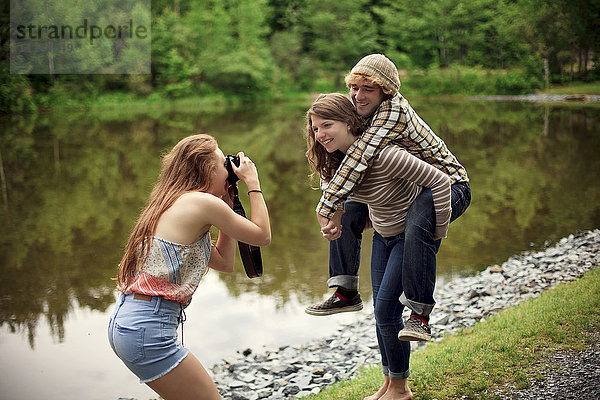Mädchen fotografiert Freund  der seinen Freund in der Nähe eines Sees huckepack trägt