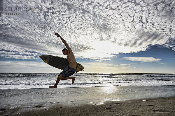 Kaukasischer Mann springt mit Surfbrett am Strand