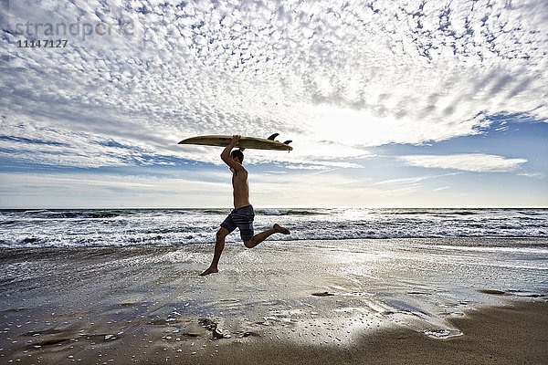 Kaukasischer Mann springt mit Surfbrett am Strand