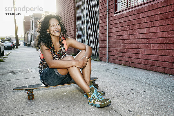 Hispanische Frau sitzt auf einem Skateboard auf einer Straße in der Stadt