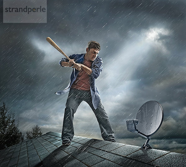 Kaukasischer Mann schlägt mit Baseballschläger auf Satellitenschüssel auf dem Dach