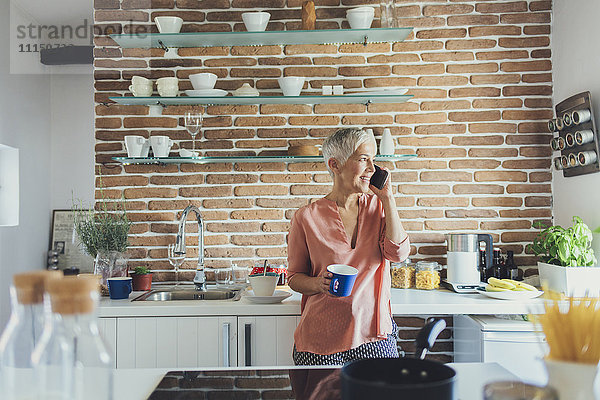 Ältere kaukasische Frau  die in der Küche mit einem Handy telefoniert