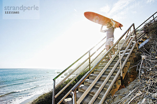 Kaukasischer Mann trägt Surfbrett auf einer Treppe am Strand