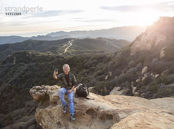 Älterer kaukasischer Mann fotografiert mit seinem Handy auf einer felsigen Bergkuppe