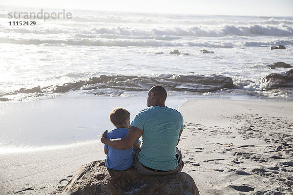 Gemischtrassiger Vater und Sohn sitzen am Strand