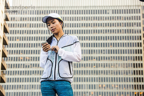 Asiatischer Läufer benutzt Mobiltelefon in der Nähe eines Hochhauses