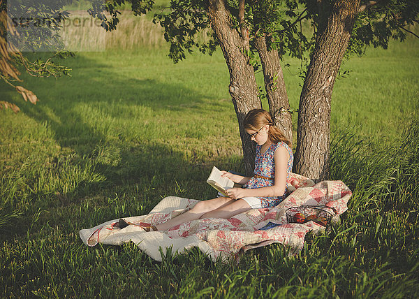 Kaukasisches Mädchen  das unter einem Baum auf einem Feld ein Buch liest