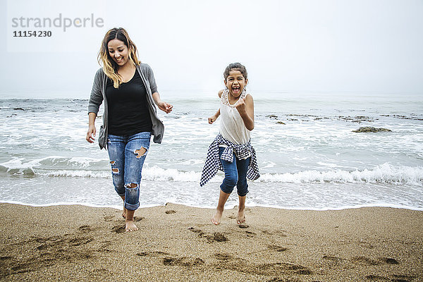 Hispanische Mutter und Tochter gehen barfuß am Strand