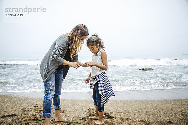 Hispanische Mutter und Tochter stehen barfuß am Strand